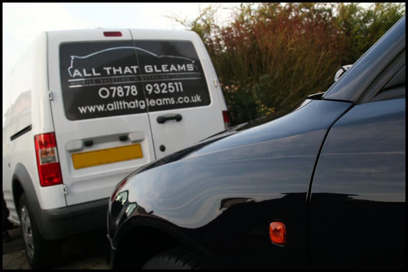 A white van with "All That Gleams" branding is parked near a dark car. The van advertises a website and contact number, promoting car valeting services and paint protection film (ppf).