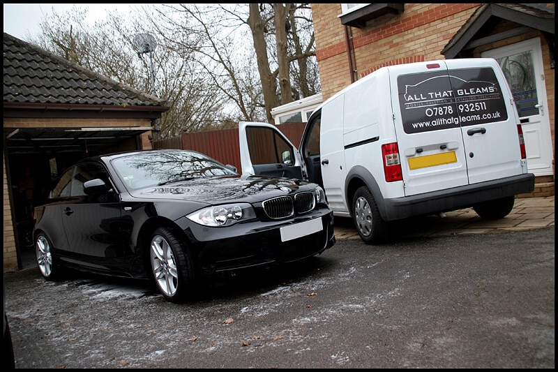 A black car fitted with paint protection film is parked beside a white van with the signage "All That Gleams" and contact details next to a brick house.