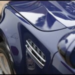 Close-up view of a blue car's left front fender, featuring a chrome vent detail and part of the mirror. Reflections are visible on the glossy surface enhanced by ceramic coating.