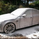 A car covered in white foam sits on a brick driveway, showcasing the meticulous care of expert car detailing. Surrounding greenery adds a touch of nature's tranquility to the scene.