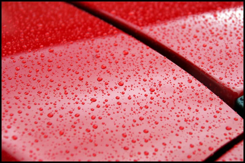 Close-up of water droplets on a red car surface, highlighting the precision of the ceramic coating and a visible gap between two panels.