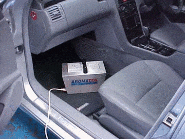 A silver box labeled "Aromatek" is placed on the passenger floor of a car with a cable connected to it. The car's interior includes two gray seats and a black dashboard, recently detailed by a professional car valeting service.