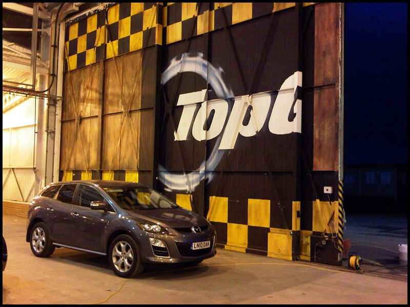 A dark SUV, sporting "Top G" on the door surrounded by a tire mark pattern, is parked in front of a large industrial building. The vehicle gleams under artificial lighting thanks to expert car detailing.