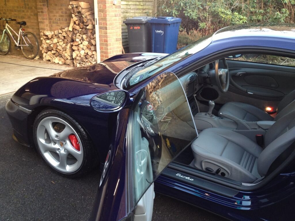 A dark blue sports car, recently treated with ceramic coating, is parked in a driveway with the driver's door open. Wood logs and a bicycle are stored in a nearby garage, while a blue recycling bin is placed outside.