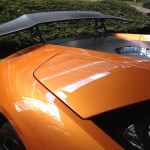 Close-up view of the rear of an orange sports car with a large black rear wing, parked near a garden area after receiving ceramic coating.