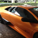 An orange sports car with black accents, freshly detailed and possibly treated with ceramic coating, is parked on a brick pavement near a staircase and greenery.