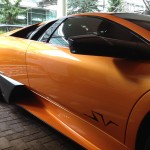 An orange sports car with paint protection film parked on a brick driveway near a modern building with greenery surrounding it.