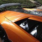 Close-up of an orange sports car with a black rear wing and a glass engine cover, parked on a brick driveway next to a small garden, highlighting its pristine condition thanks to professional car valeting.