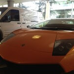 An orange Lamborghini sports car is parked next to a white van with "All That Gleams" branding in a shaded area, highlighting expert car detailing services.