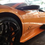 Close-up view of the side of an orange sports car with black wheels and "SV" lettering on the door, parked on a brick surface, showcasing its pristine finish thanks to high-quality paint protection film.