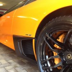 Close-up of an orange sports car parked, focusing on the left front wheel and fender. The vehicle boasts black rims and a low profile with sleek, aerodynamic lines enhanced by meticulous car detailing.