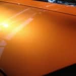Close-up of a shiny orange car hood with a reflection of the sun and surrounding structures, showcasing the smooth finish provided by paint protection film.