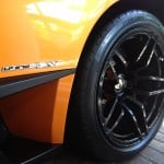 Close-up of the rear wheel of an orange sports car with "LP 670-4 SV" labeling, showcasing black alloy rims and performance tires on a stone-paved surface. The high-quality finish reflects precision car detailing.