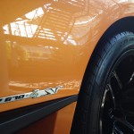 Close-up of an orange sports car showing a "LP 670-4 SV" emblem and a black wheel with visible tire treads reflecting the surroundings. The vehicle's glossy finish benefits from paint protection film, ensuring it remains pristine even after rigorous drives.