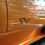 Side view of an orange sports car with a black "SV" logo on the door, parked in front of a modern building with glass windows, showcasing its sleek design enhanced by meticulous car detailing.