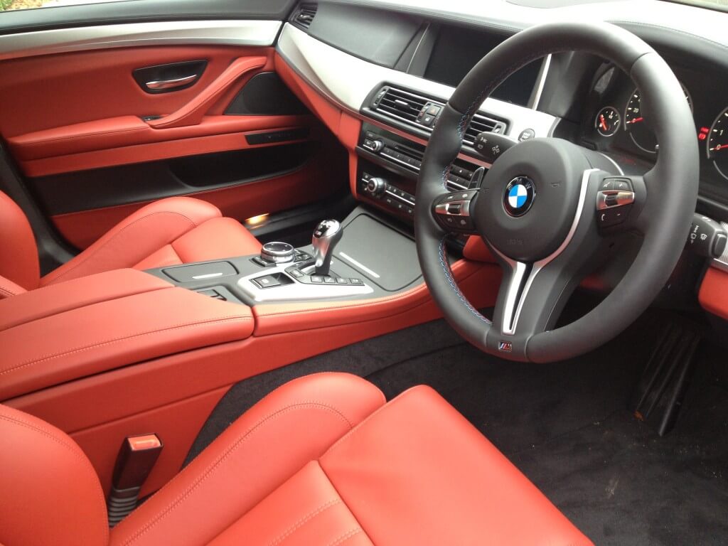 The image shows the interior of a luxury car with red leather seats, a black dashboard, steering wheel, and various control buttons. It appears to be the interior of a BMW vehicle that has undergone meticulous car detailing for an immaculate finish.
