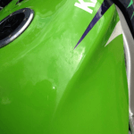 Close-up of a green motorcycle fuel tank showing part of the brand name "Kawasaki" with a reflection of the sky, protected by ceramic coating.