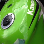 Close-up of a green Kawasaki motorcycle's fuel tank, showing the brand logo and fuel cap. The tank and surrounding parts exhibit signs of use and wear, but the addition of ceramic coating has helped maintain a sleek appearance.