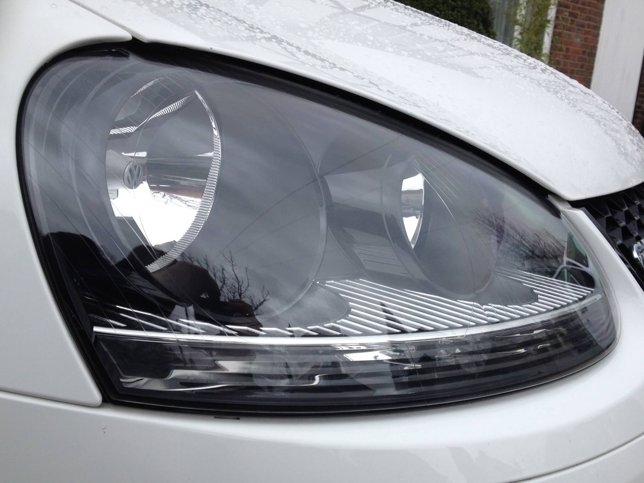 Close-up view of a car's headlight with a clear lens and sleek design, showing reflections and minor rain droplets on the surface, enhanced by paint protection film (PPF).