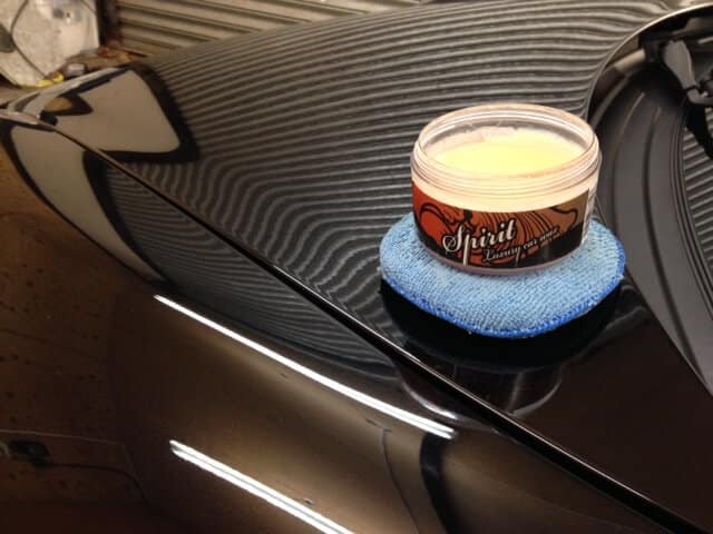 A container of car wax labeled "Spirit Luxury" placed on top of a blue polishing pad atop the hood of a meticulously polished, black vehicle receiving expert car valeting.