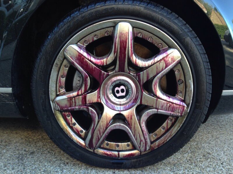 Close-up of a car wheel with a metallic rim stained by a red cleaning solution, mounted on a tire. The center of the rim displays a "B" logo from recent car detailing. The visible tire treads rest against the ground surface.