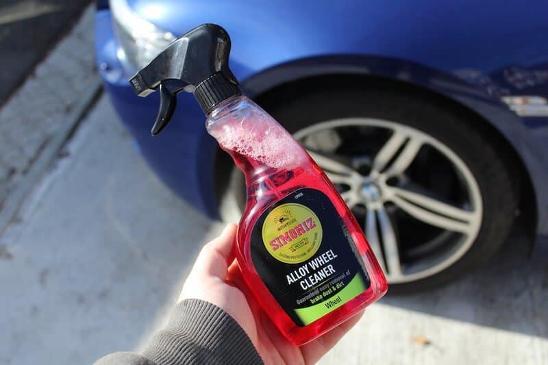 A hand holding a bottle of Simoniz Alloy Wheel Cleaner in front of a car wheel, perfect for car detailing. The cleaner is in a clear bottle with red liquid and a spray nozzle.