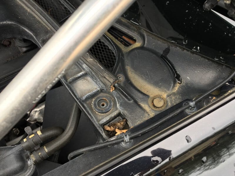 Close-up of a car engine compartment with metal parts, a wire, and some leaves trapped near the edge, highlighting the effectiveness of car detailing.