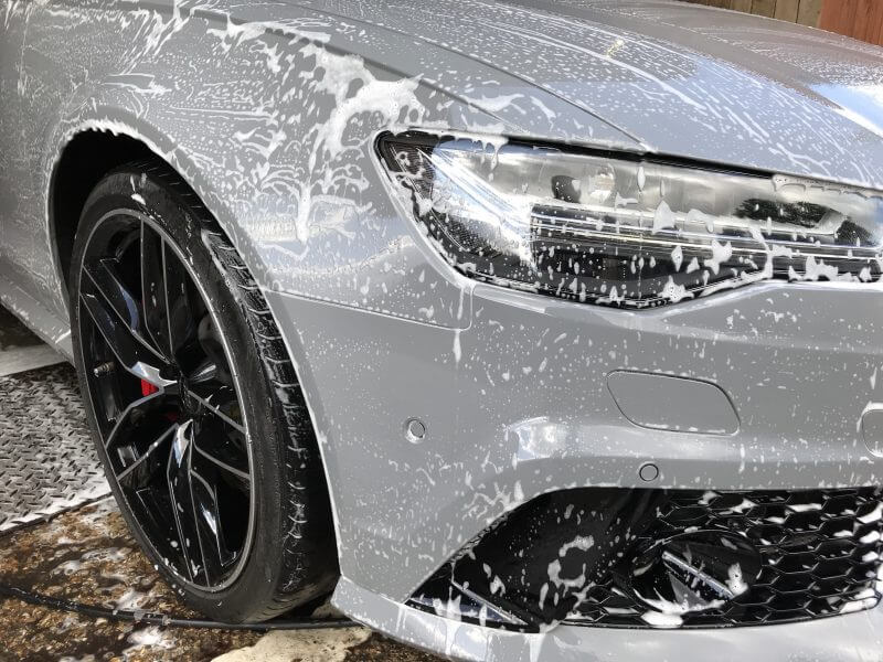 A gray car with black wheels, covered in soap suds, is undergoing car detailing, focusing on the front headlight and wheel area.