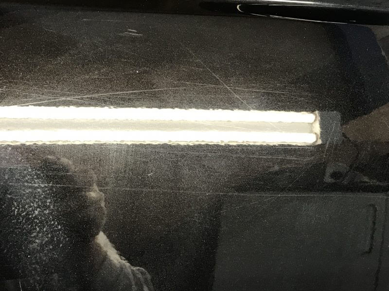 Close-up of a dark, scratched surface with a rectangular cutout containing two parallel strips of white material. The surface shows signs of wear, ideal for car detailing or paint protection film application. Reflections are partially visible on the surface.