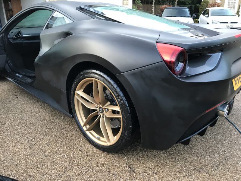 A matte black sports car with gold alloy wheels is parked on a driveway, showcasing expert car detailing. The driver's side door is open.