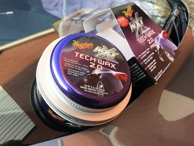 A container of Meguiar's NXT Tech Wax 2.0 sits on a car hood next to its open packaging, which features product details and an image of a car, highlighting its synergy with ceramic coating.