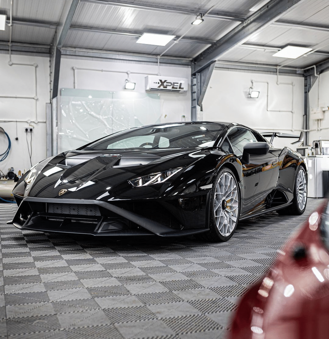 A sleek black Lamborghini sports car is parked in a well-lit, industrial-style garage with white walls and a checkered floor, showcasing its premium car detailing and paint protection film.