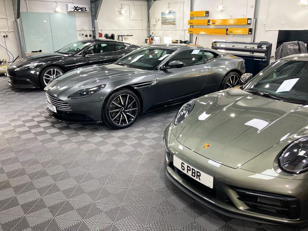Three luxury sports cars are parked inside a well-lit garage with a checkered floor. Two grey Aston Martins gleam beside a silver Porsche, proudly displaying its "6 PBR" license plate. The glossy finishes look impeccable, hinting at recent car valeting and ppf treatments.