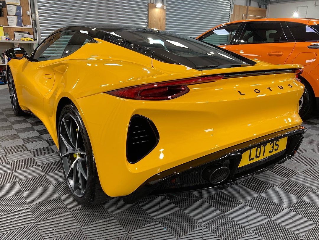 A bright yellow Lotus sports car is parked indoors on a patterned floor, showcasing its license plate "LOT 35". A glimpse of an orange vehicle is seen nearby, suggesting a detailed car valeting session.