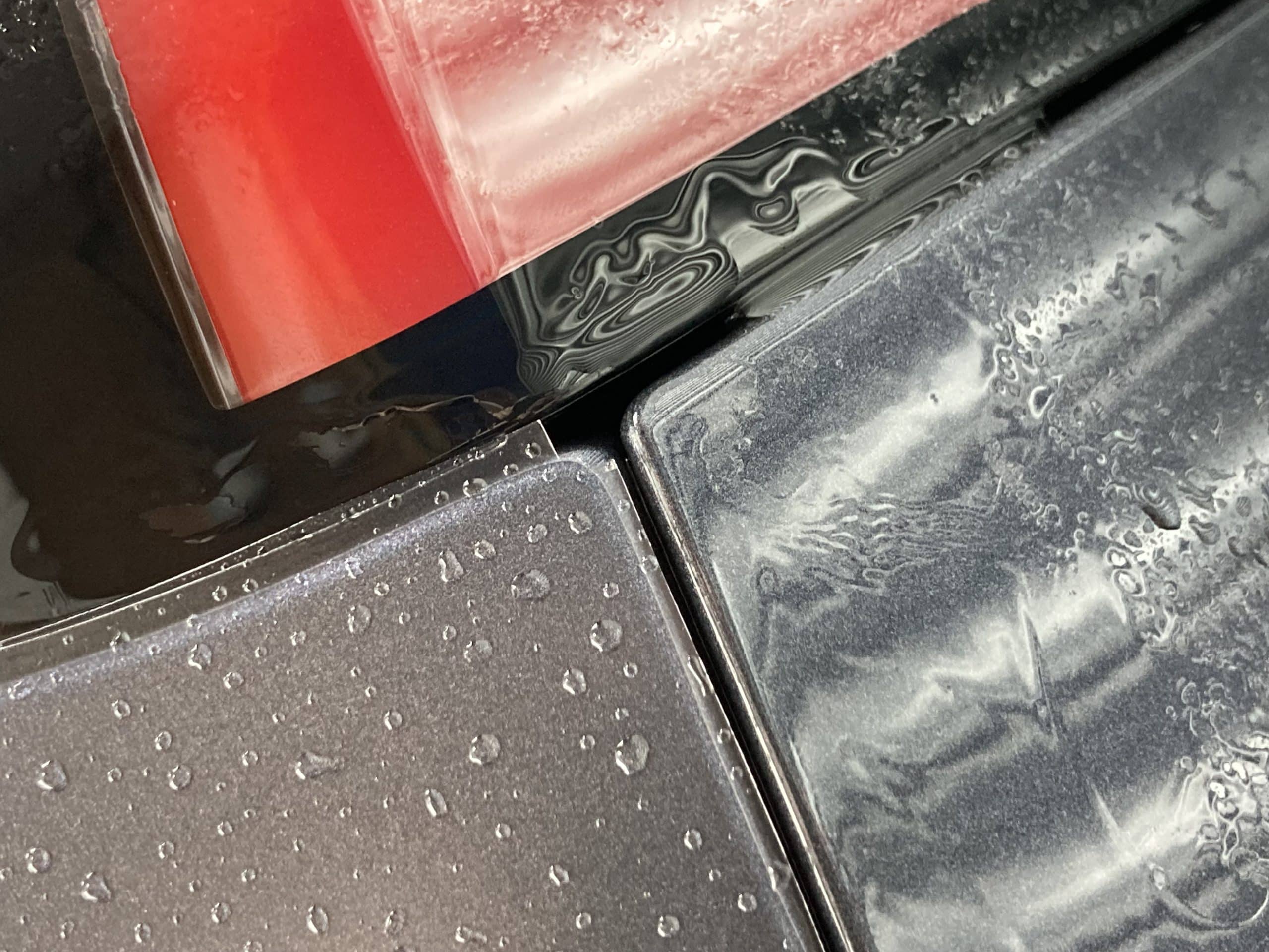 Close-up of water droplets on the surfaces of overlapping sheets of glossy material, with shades of red, black, and gray, showcasing the precision of ceramic coating in car detailing.