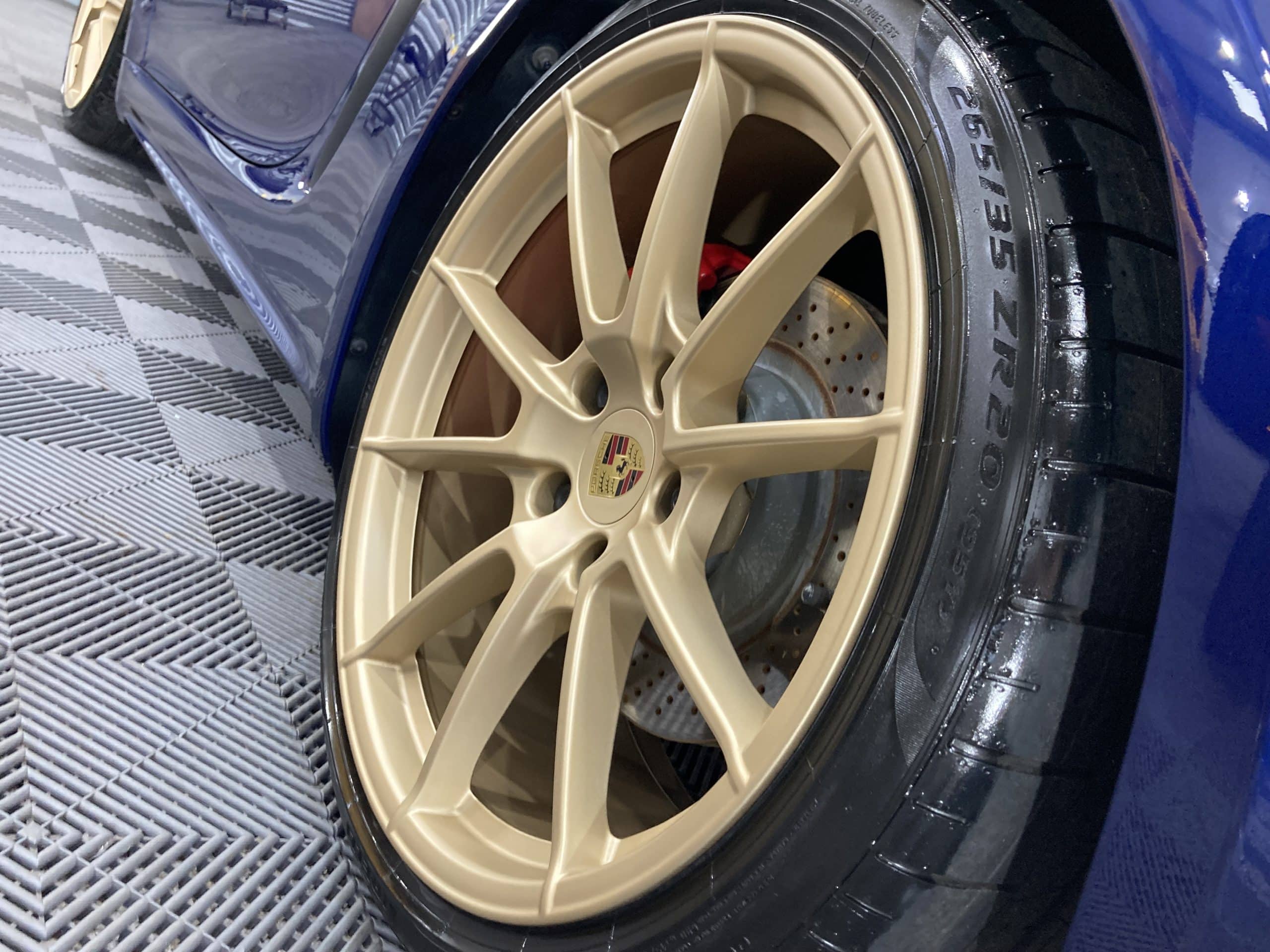A close-up of a gold alloy wheel on a blue car, displaying the tire size 265/35 ZR20 and a visible disc brake with a Porsche logo on the hub. The car detailing highlights the shine, enhanced by paint protection film for added durability.