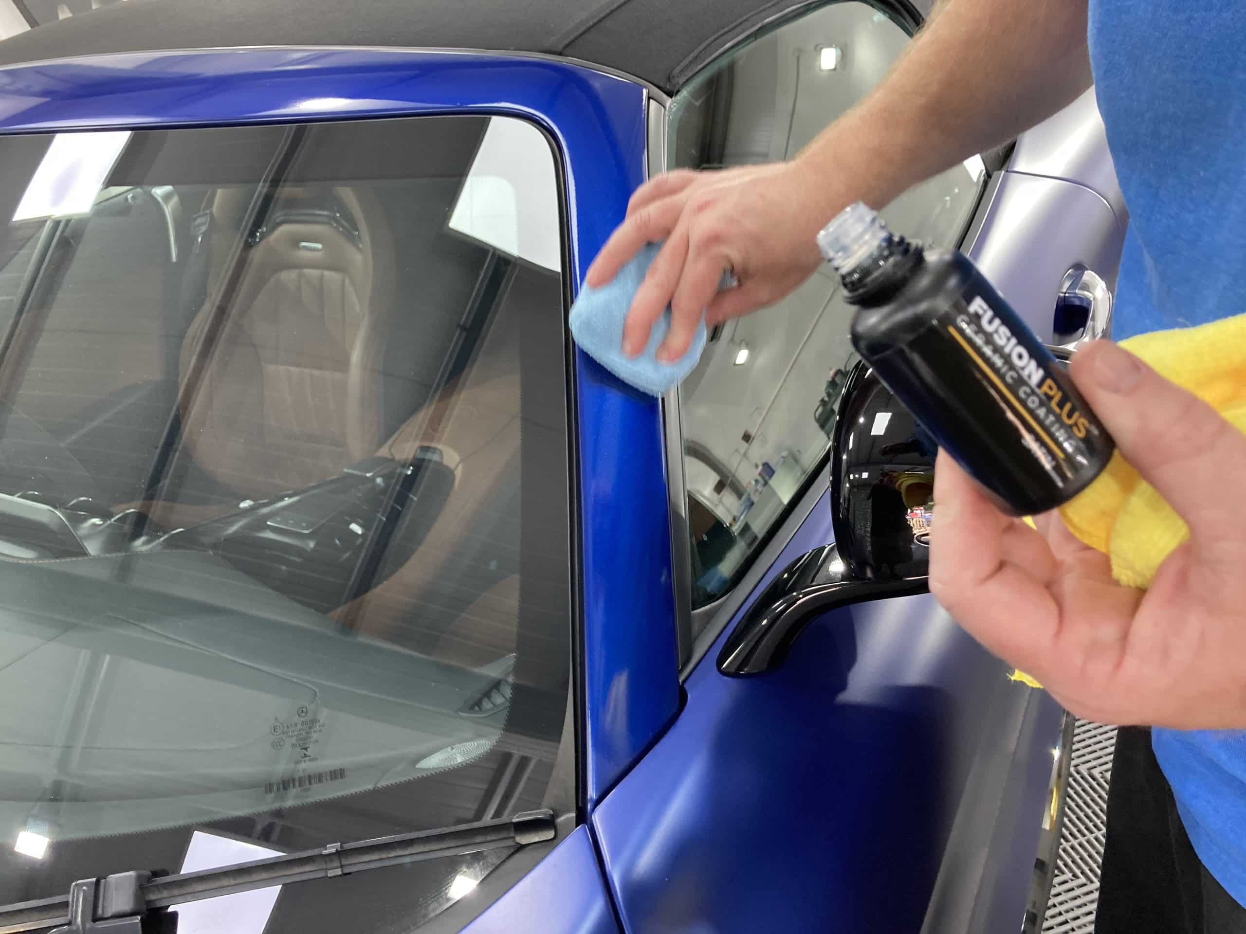 A person applies Fusion Plus ceramic coating to the windshield of a blue car using a blue cloth to enhance its paint protection film.