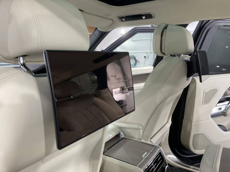 The image shows the interior of a luxury car with beige leather seats, enhanced by ceramic coating. Two screens are mounted on the back of the front headrests, facing the rear seats. The car door is open, revealing the rear area.
