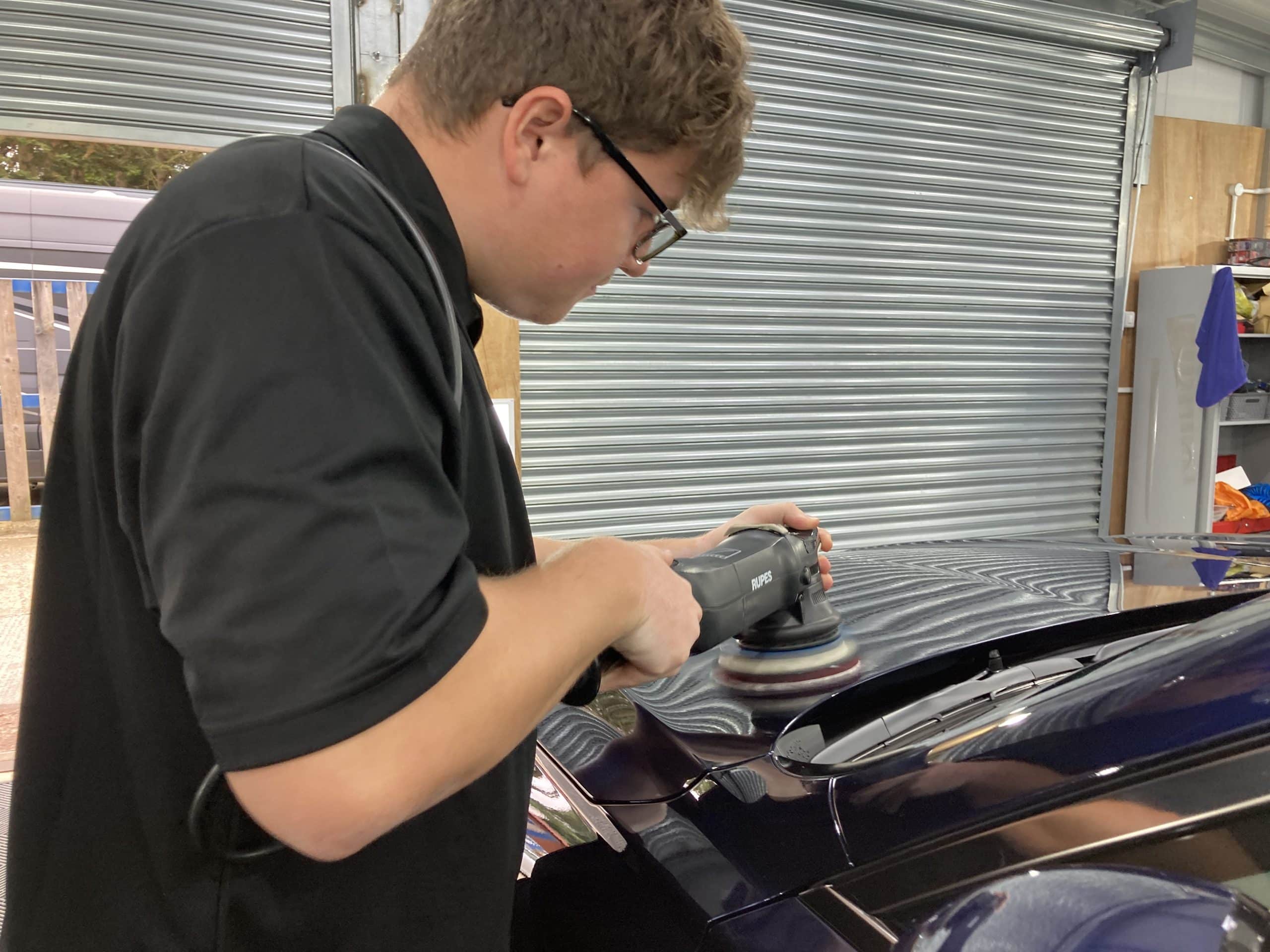 A person in a black shirt is performing car detailing using an electric polisher on the hood of a dark-colored car inside a workshop with metal roll-up doors.