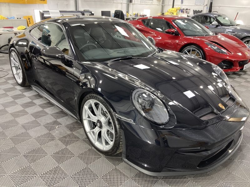 A black Porsche sports car parked indoors on a checkered floor, featuring paint protection film to maintain its sleek look. A red car is also visible.