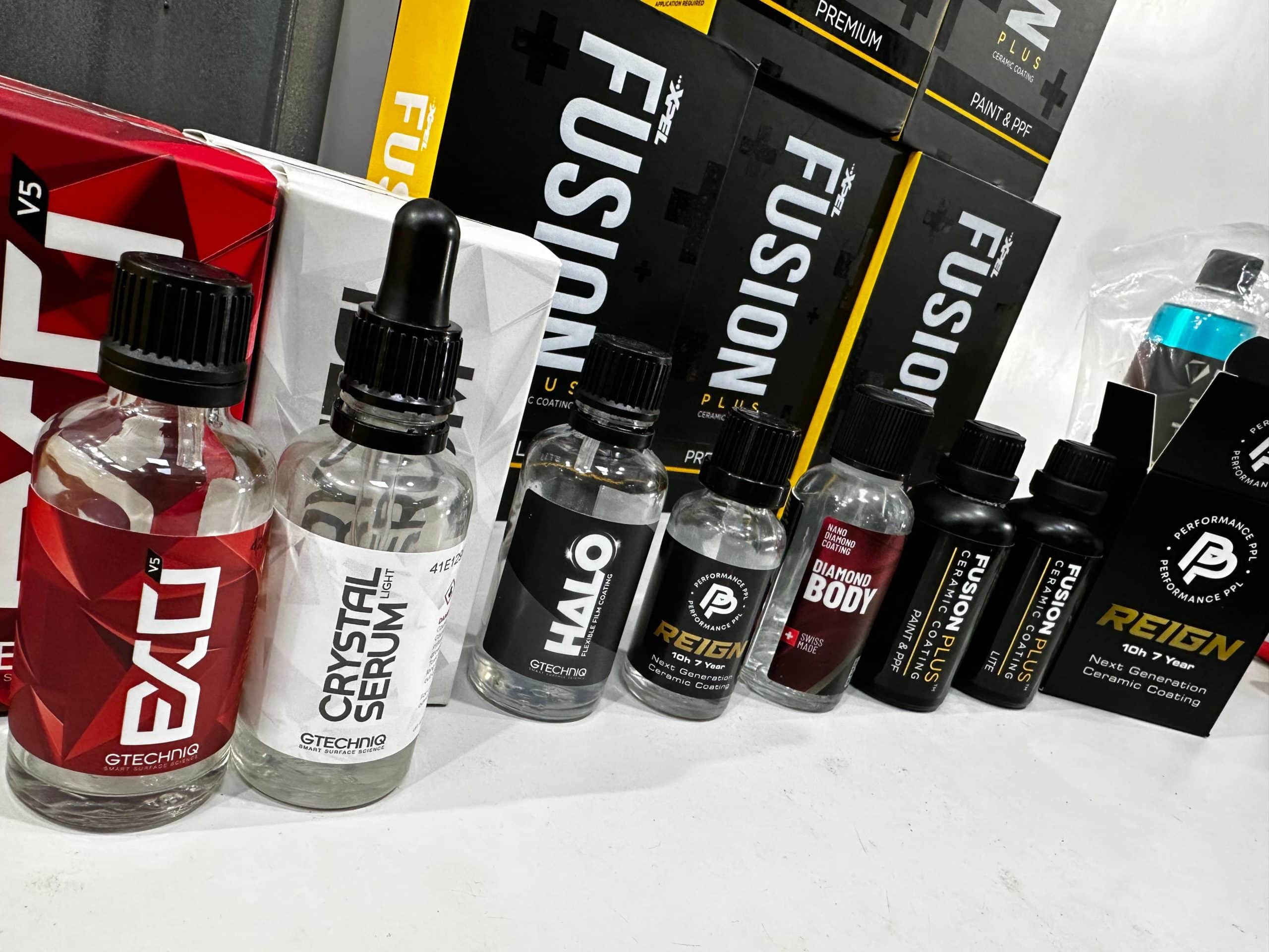 A line-up of various car care products including bottles of ceramic coatings, sealants, and cleaning agents on a white surface with branded packaging. The display offers essential items for car valeting enthusiasts seeking top-notch finish and protection.