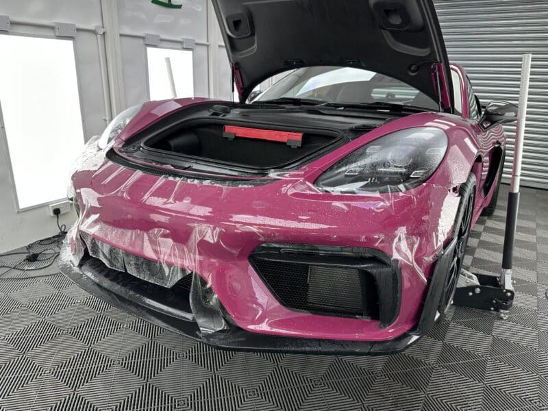 A pink car with an open hood is being worked on in a garage, where the front bumper is partially covered with protective film. Curious about how much PPF costs? The team carefully applies it, ensuring every detail enhances the vehicle's sleek appearance.