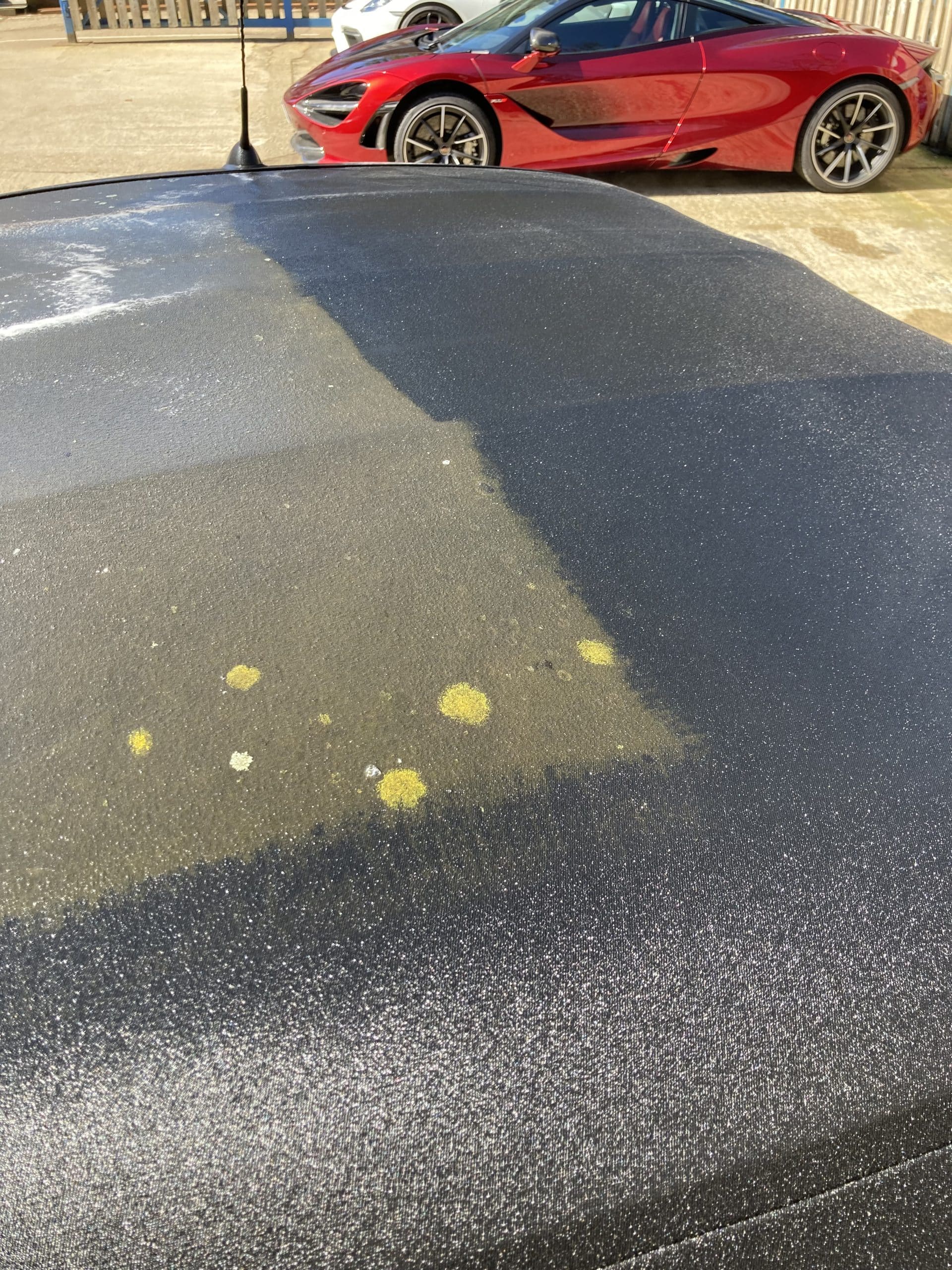 The image shows the roof of a black car with mossy or moldy spots on it. Parked next to it is a red sports car, highlighted by its spotless finish thanks to paint protection film.