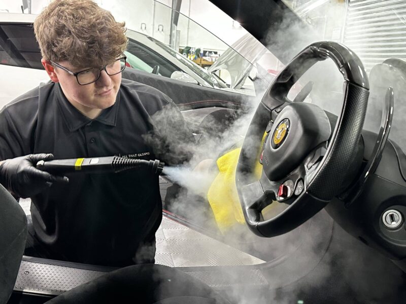 A person wearing gloves and a black shirt uses a steam cleaner on the steering wheel of a car, with steam visibly rising. The car's interior appears to be undergoing detailing and car valeting in a garage.