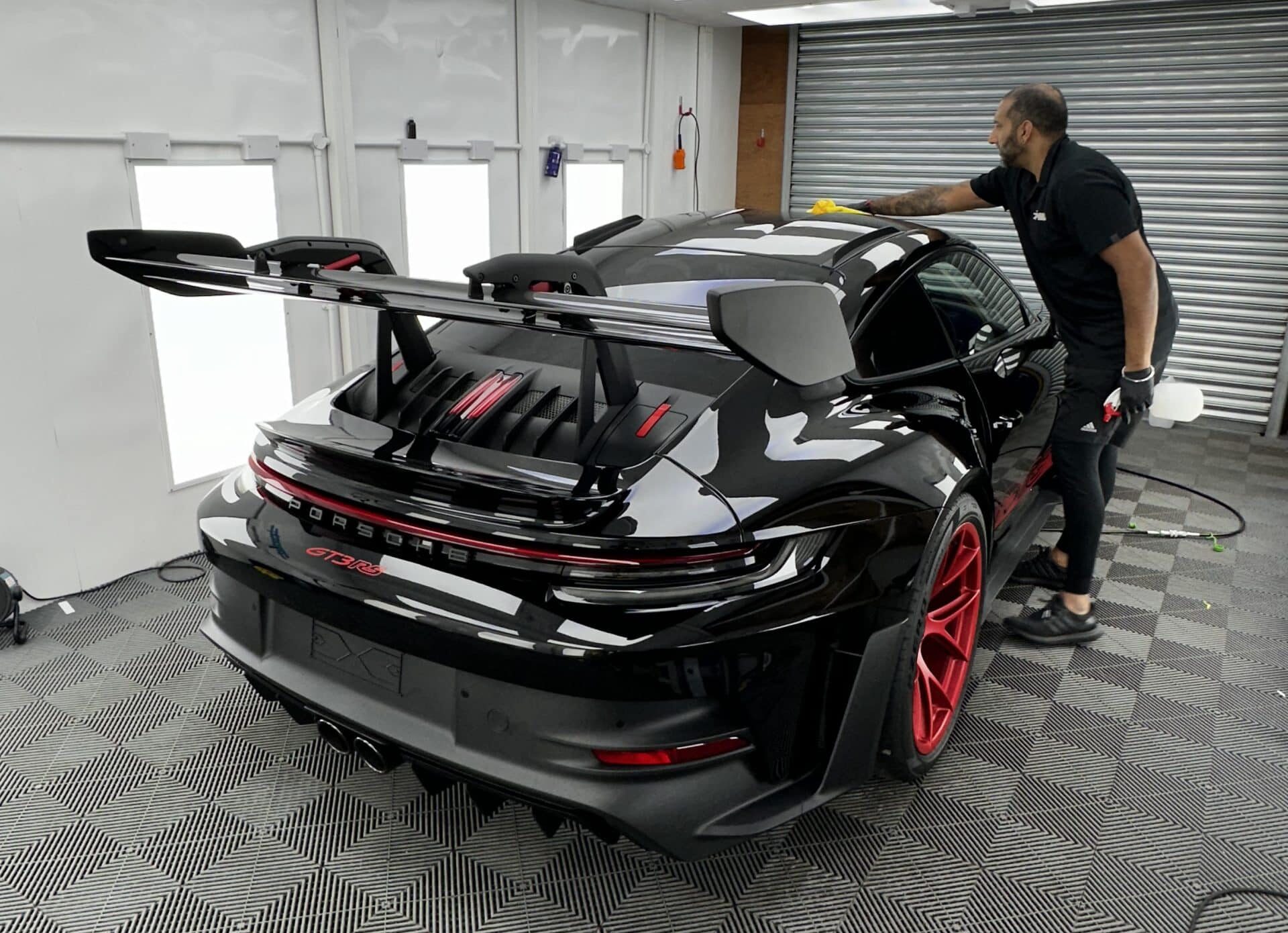 A person is car detailing a black sports car with red accents and a large rear spoiler in a well-lit garage, applying paint protection film for an immaculate finish.